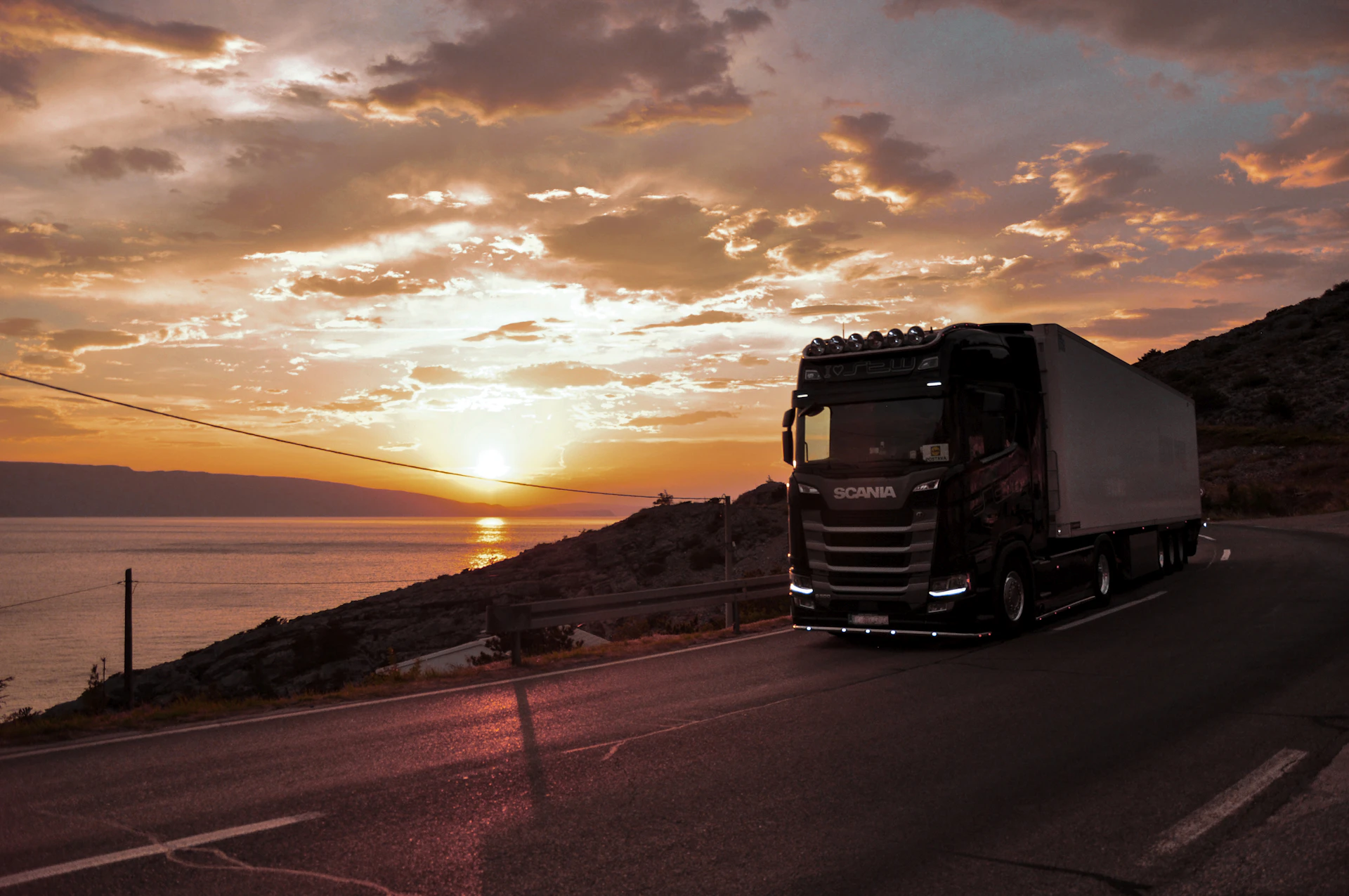Modern semi-truck driving along a coastal road at sunset