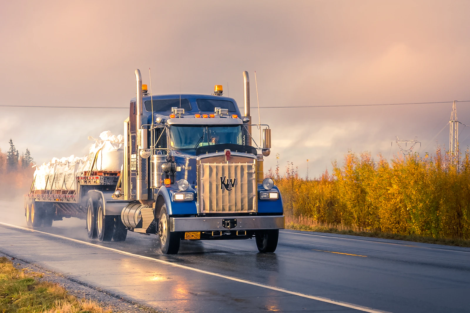 Kenworth semi-truck hauling a flatbed load at sunset on a wet road